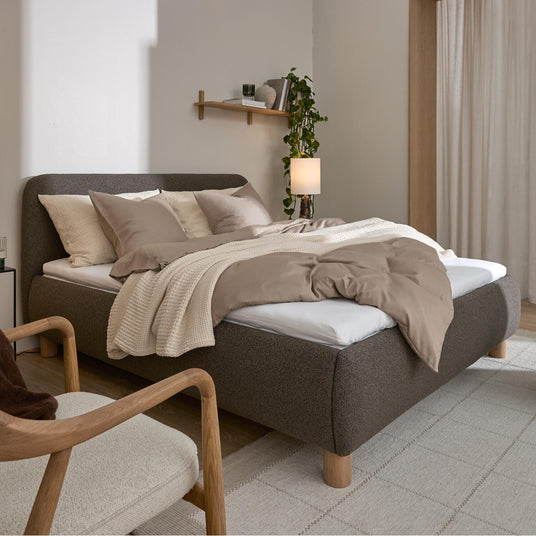 Soveværelse med polstret grå seng, beige sengetøj og strikket plaid, lyse gardiner og væghylde med plante og lampe i blødt dagslys.