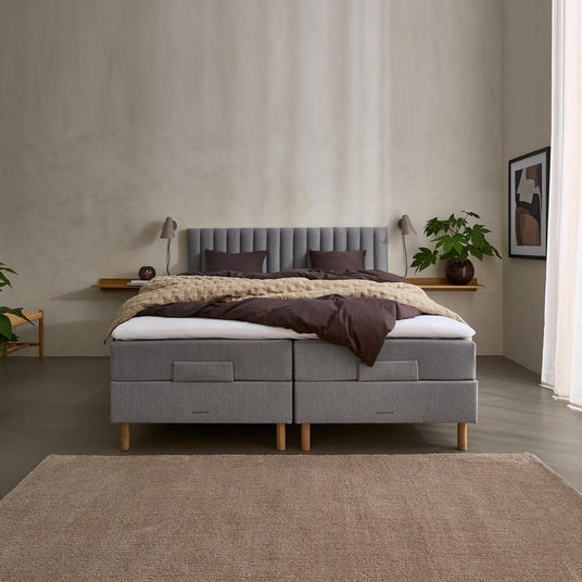Grå polstret seng i et minimalistisk soveværelse med brunt sengetøj, væglamper, plante, vægkunst og beige tæppe i blødt dagslys.