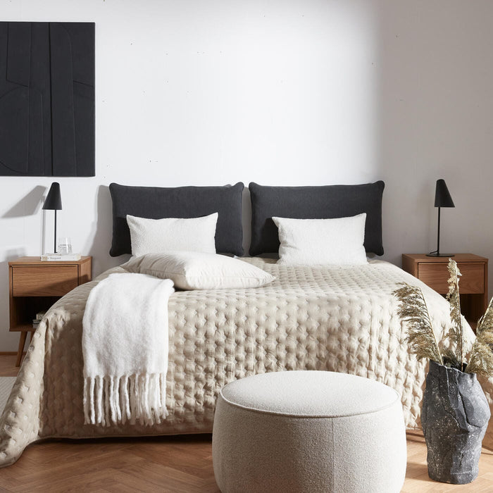 Redt dobbeltseng i beige med hvide puder og plaid, sorte sengelamper og natborde i træ; rund puf og vase med tørrede strå i et lyst soveværelse, bogen Voyage på bordet.
