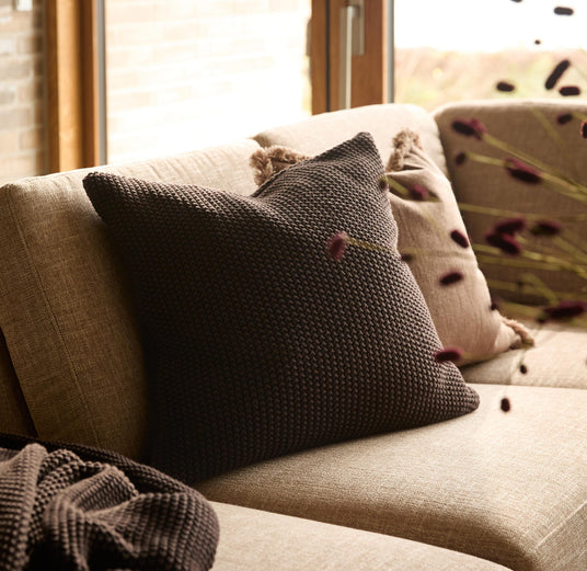Beige sofa med strikket mørkebrun pude, lys pude og matchende plaid i blødt dagslys ved vinduet.