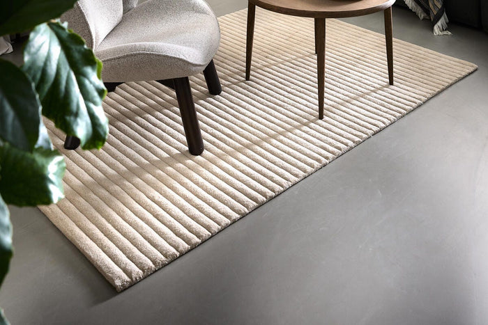 Lyst rillet tæppe i stue med rundt sofabord i træ, grøn sofa med plaid og skulpturel lænestol.