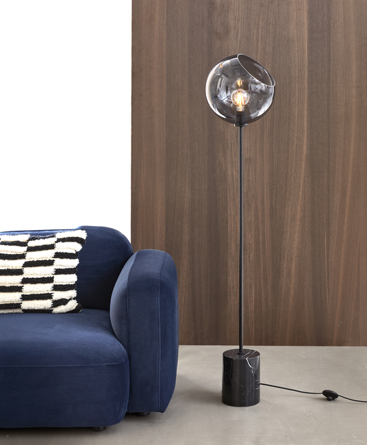 Gulvlampe med røgfarvet glaskupple og sort marmorfod ved siden af en mørkeblå sofa med sort‑hvid stribet pude, foran træpanel og betongulv.