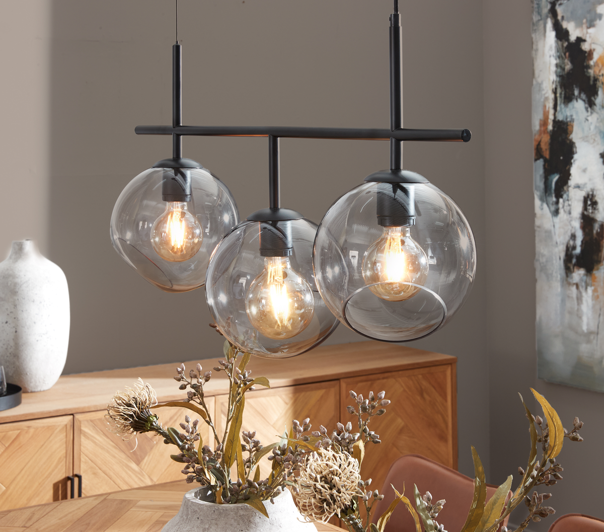 Trelampe-pendel med røgfarvede glaskupler over et spisebord, tændte pærer og en træskænk i baggrunden.