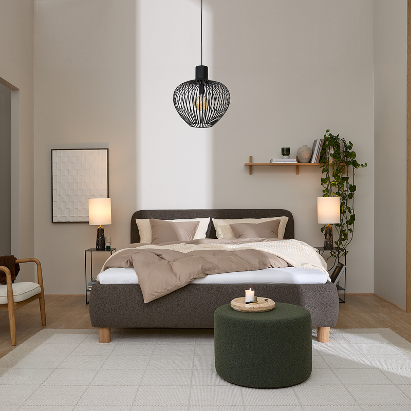 Lyst soveværelse med mørkegrå polstret seng og beige sengetøj, sort trådpendel i loftet, to bordlamper, væghylde med hængeplante og bøger samt grøn rund puff med bakke og tændt lys på lyst tæppe.