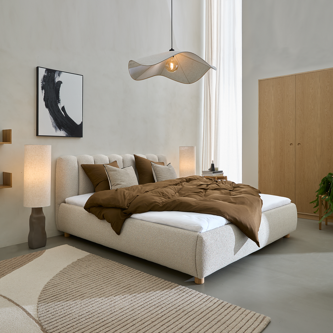 Lyst soveværelse med beige polstret seng, brun dyne, skulpturel pendel, to gulvlamper, lyse gardiner, træskab og beige mønstret tæppe.
