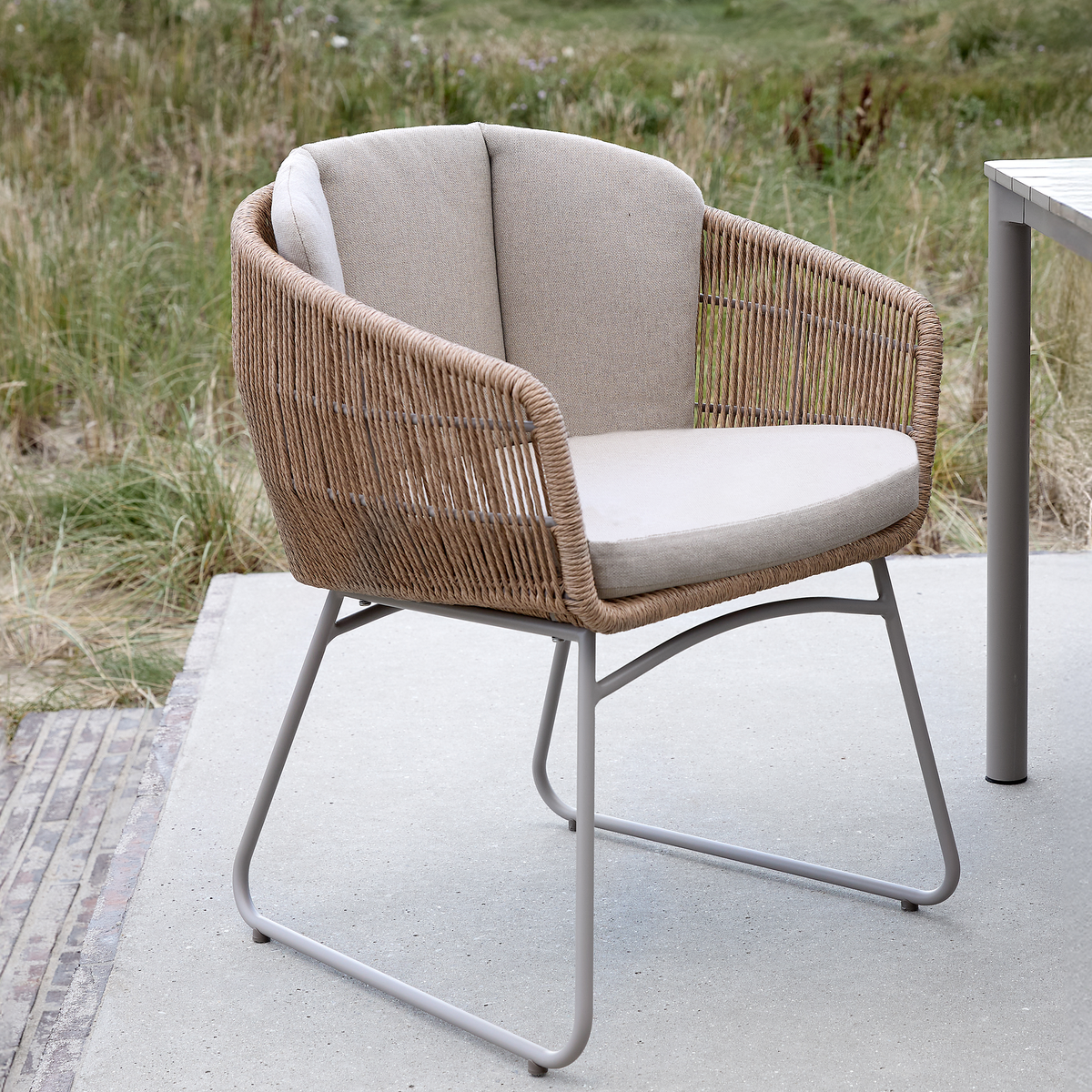 Udendørs stol i flettet reb/rotting-look med beige hynder og gråt metalstel på terrasse ved siden af et bord.