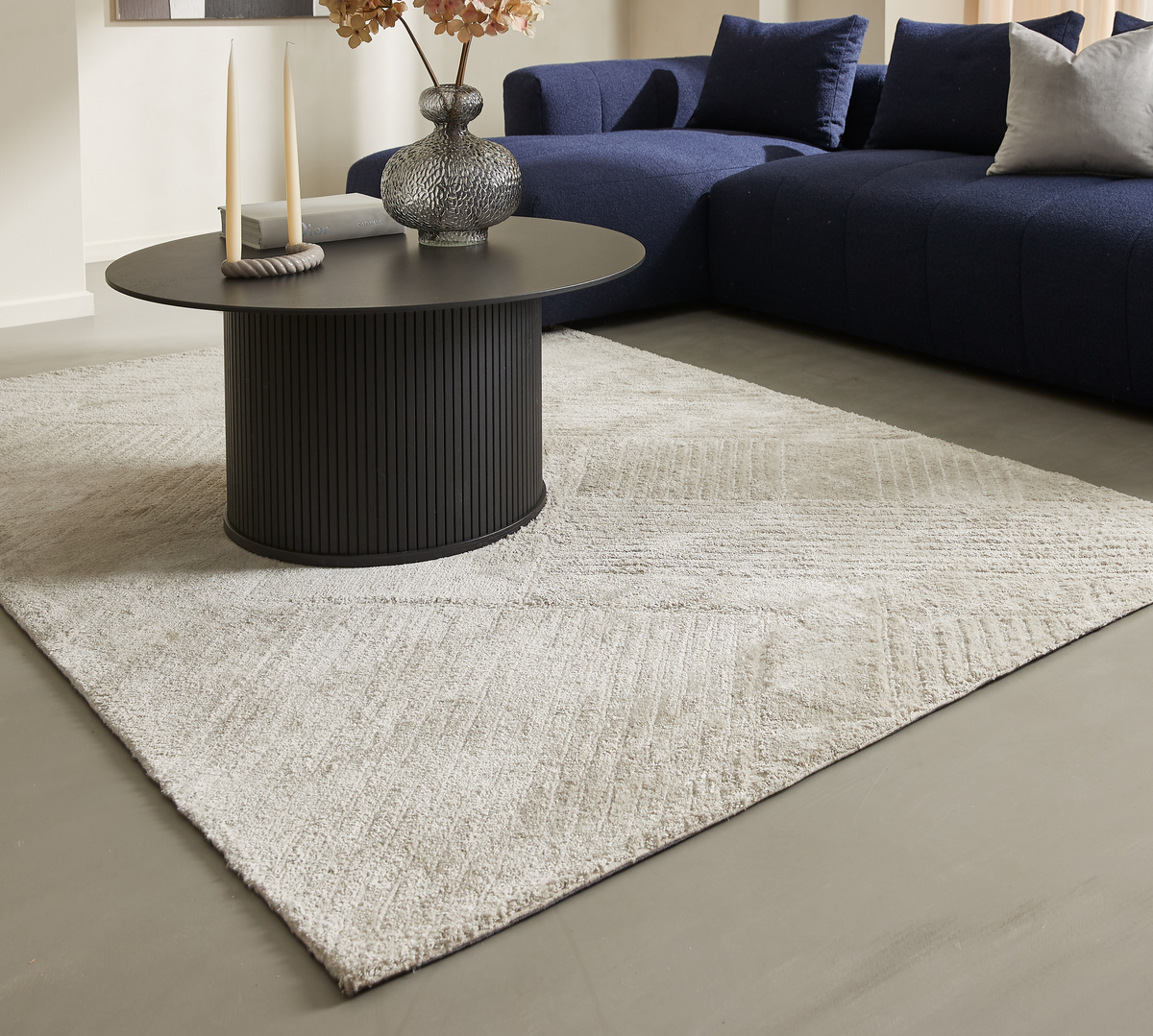 Mørkt rundt sofabord på lys beige tæppe med diskret mønster, ved siden af blå modulsofa med puder, dekoreret med lys, bog og glasvase med blomster.