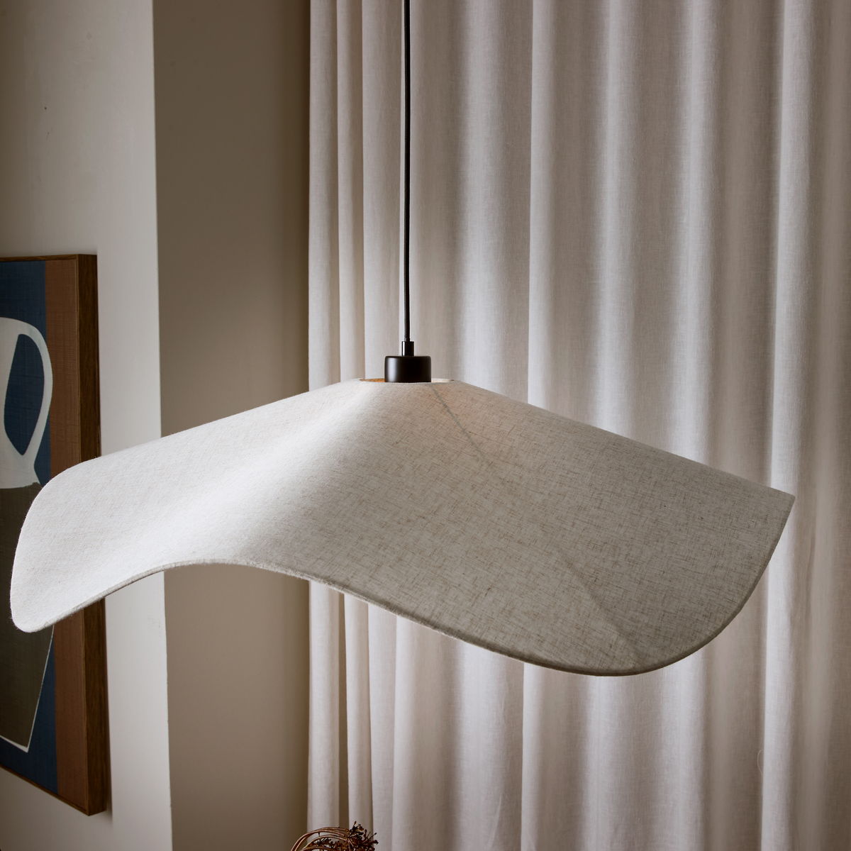 Bølgeformet pendellampe i beige tekstil hænger foran lyse gardiner i blødt dagslys.