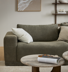 Grøn stofsofa med lyse puder i stuen, runde sofaborde med magasin og briller, hylder og vægkunst i baggrunden.