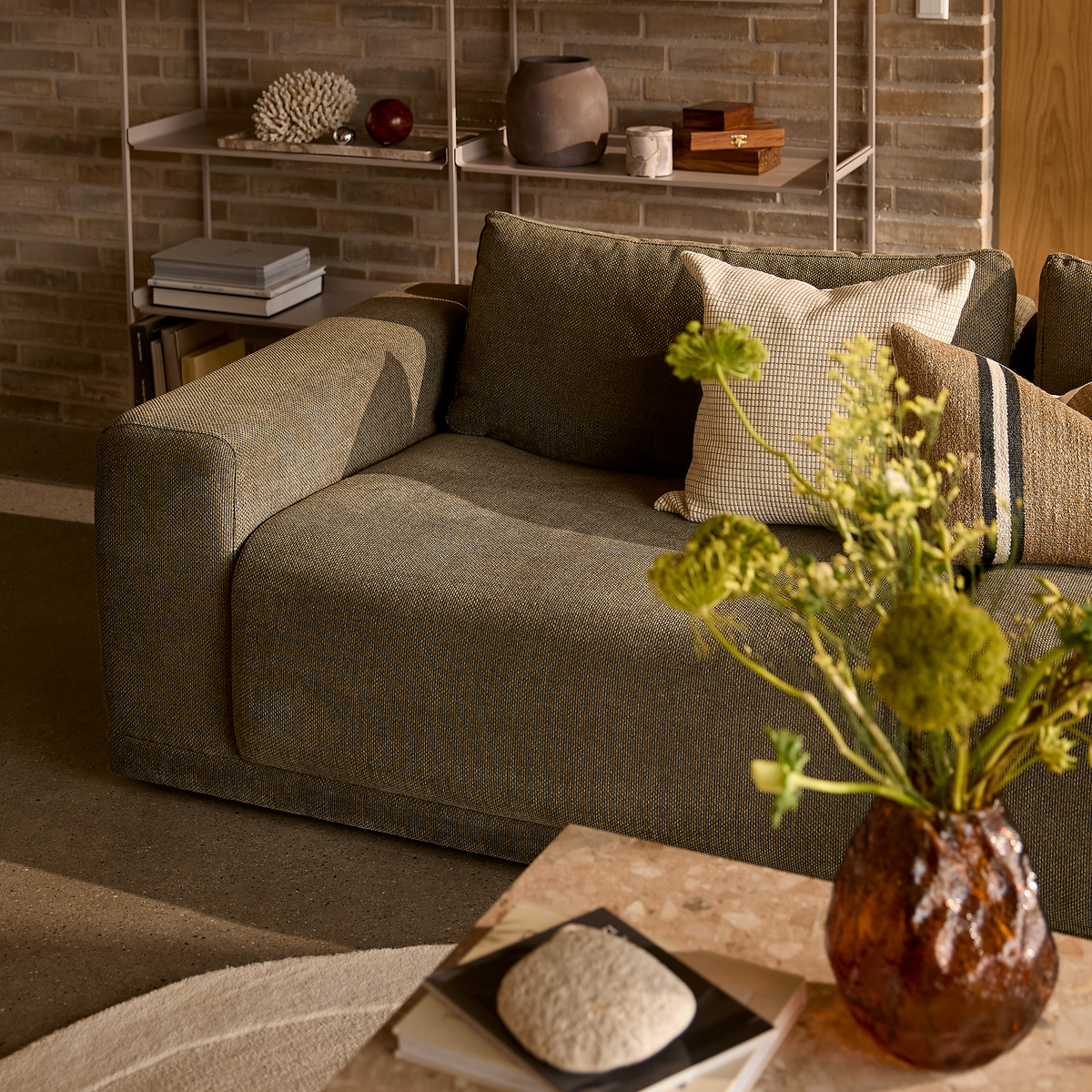 Stue med grå stofsofa, mønstrede puder og ravfarvet vase med grønne blomster på sofabord; murstensvæg og hylder i varmt dagslys.