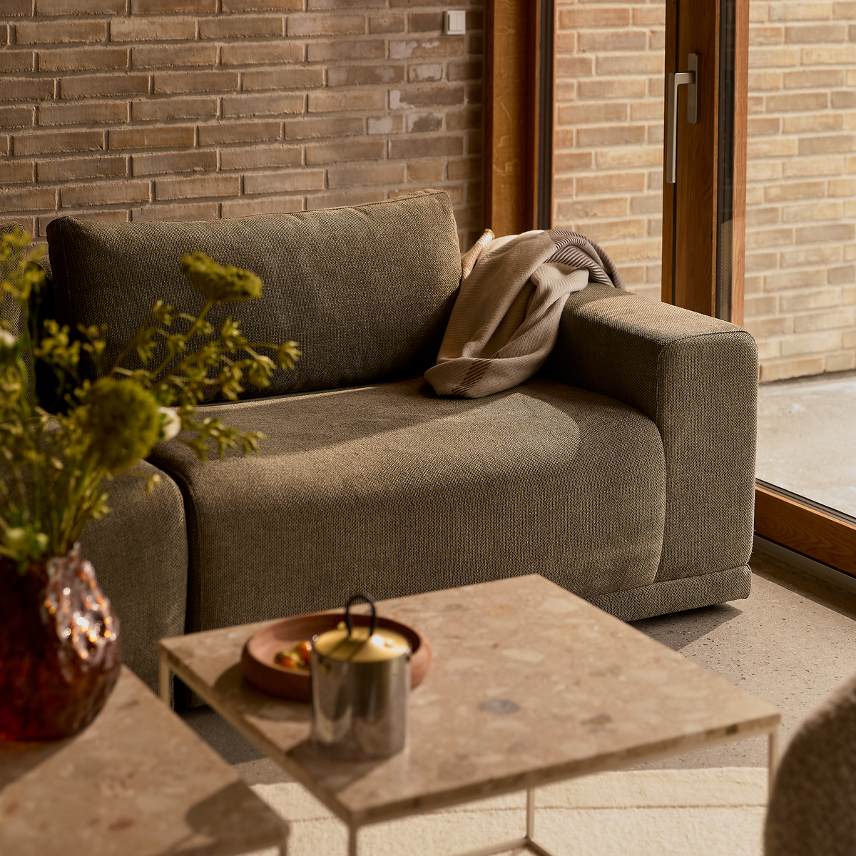 Hyggekrog i stuen med grå‑beige sofa, plaid over armlænet og to stensofaborde med vase med grønne grene og et lille fad; varmt dagslys fra glasdør mod murstensvæg.
