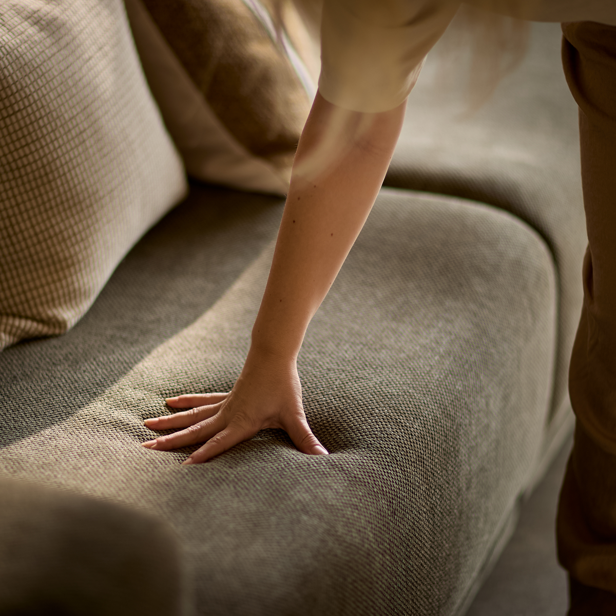 Hånd presser ned i en tekstureret beige sofasæde for at vise blødhed, puder i baggrunden.