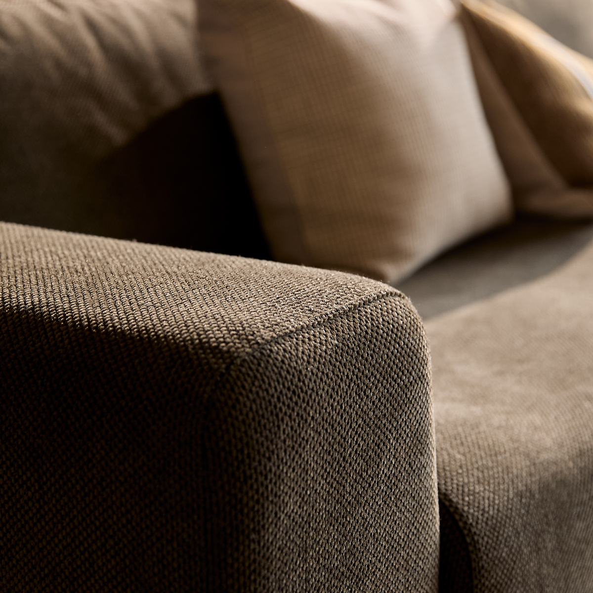 Nærbillede af brun tekstilsofa med struktureret armlæn og beige puder i varmt dagslys.