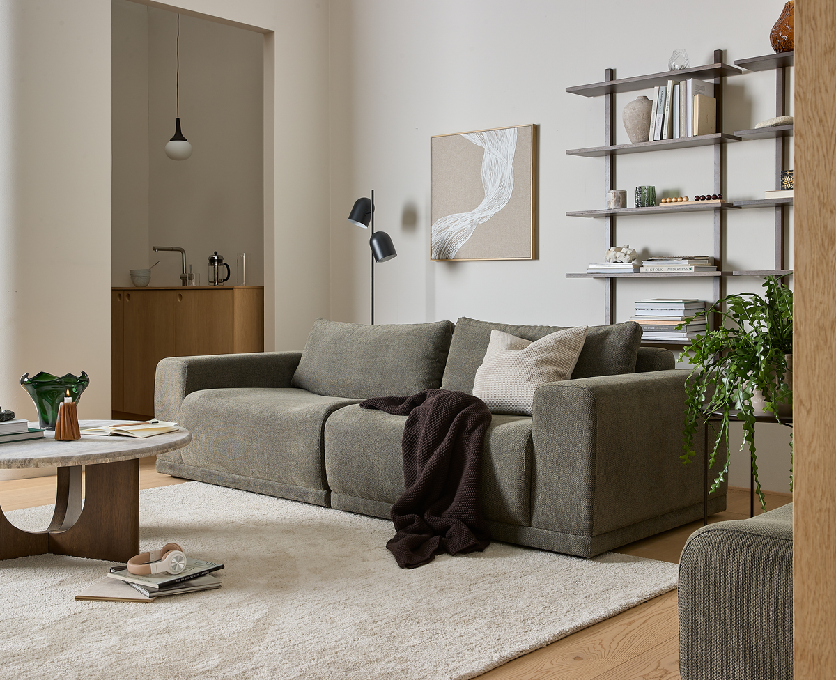 Lyst, roligt stue-miljø med grøngrå sofa, beige pude og mørkt tæppe, rundt sofabord i træ/sten, vægmonteret reol med bøger og dekorationer, gulvlampe og grøn plante på lyst tæppe.