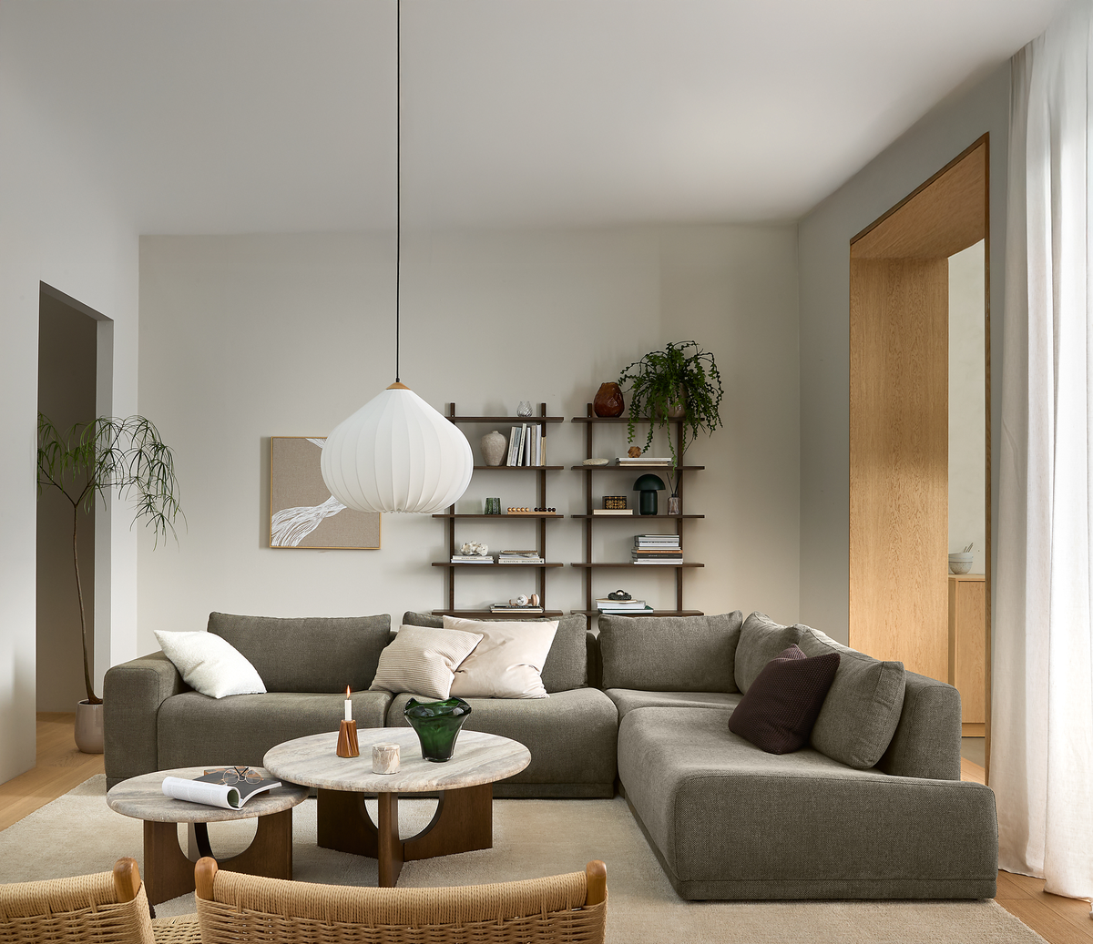 Lyst, minimalistisk stue med grå modulsofa, runde sofaborde med stenplader, hvid rispendel, åben reol med bøger og planter samt lyse gardiner.