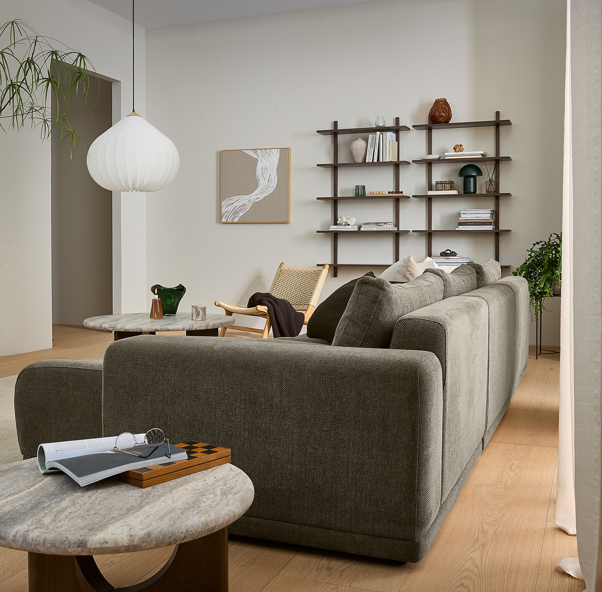Lyst, skandinavisk stue med grå modulsofa set bagfra, vægmonterede hylder med bøger og dekoration, rund hvid pendel, to runde marmorborde og grønne planter på trægulv i dagslys.