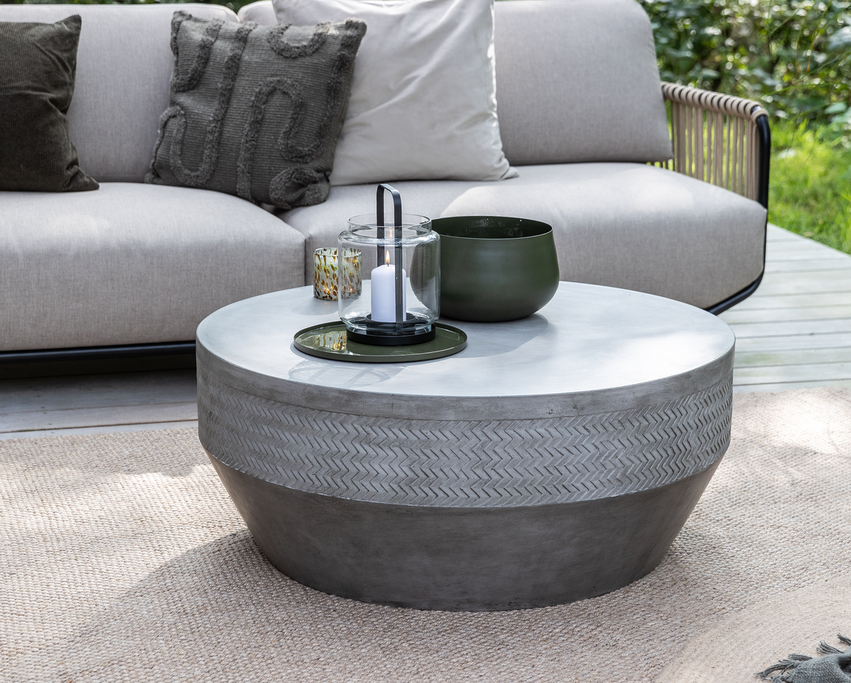 Rundt betonbord udendørs med sildebensmønster, grøn skål og glaslanterne med hvidt lys på bakke på træterrasse foran beige sofa.