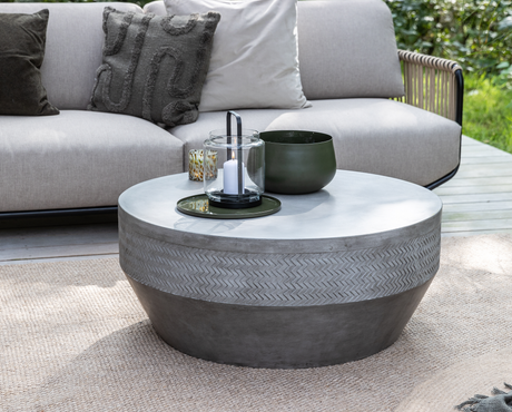 Rundt betonbord udendørs med sildebensmønster, grøn skål og glaslanterne med hvidt lys på bakke på træterrasse foran beige sofa.
