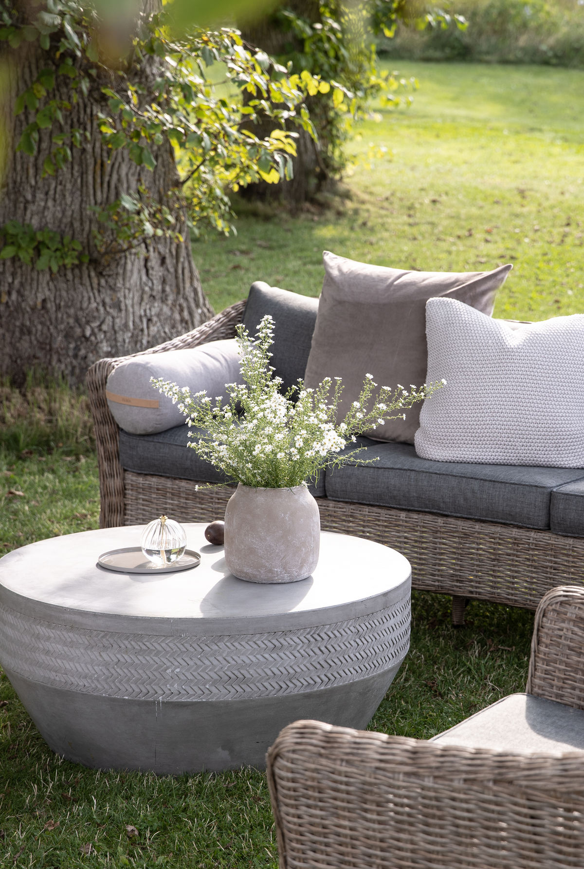 Udendørs lounge med fletsofa med grå puder og rundt betonbord med en vase med hvide blomster i en have.