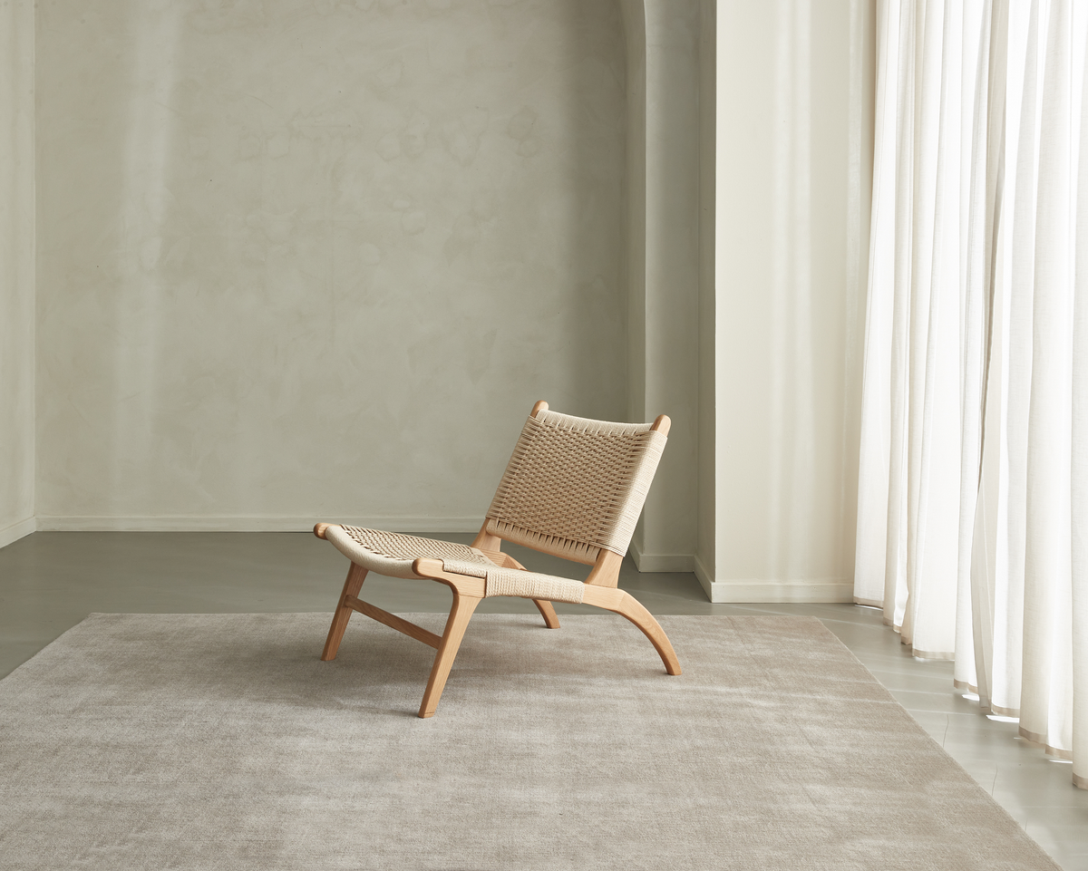Lav loungestol i lyst egetræ med flettet sæde på et beige tæppe i et lyst, minimalistisk rum med hvide gardiner.
