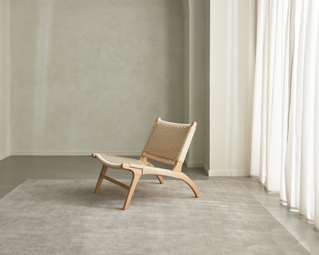 Lav loungestol i lyst egetræ med flettet sæde på et beige tæppe i et lyst, minimalistisk rum med hvide gardiner.