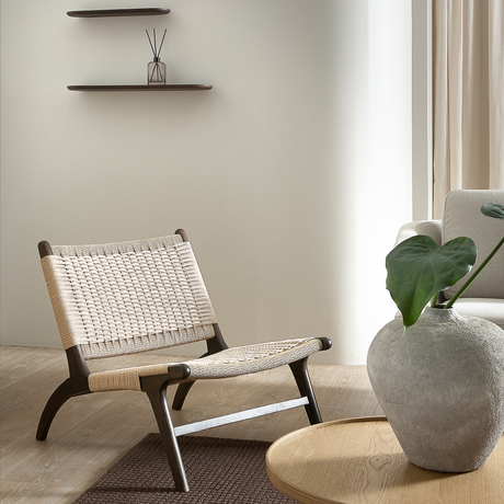 Lav loungestol med vævet sæde og mørke træben ved siden af et rundt egetræsbord med stor vase og grønt blad i en lys, minimalistisk stue.
