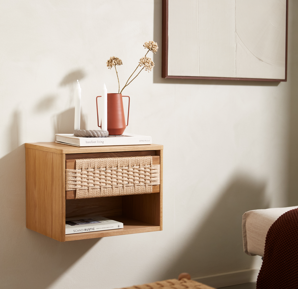 Vægmonteret natbord i eg med vævet front, terrakottakande med tørrede blomster, to hvide lys og bøgerne barefoot living og SCANDI RUSTIC.