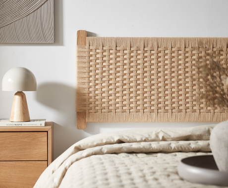 Nærbillede af lyst soveværelse med flettet sengegavl i naturmateriale, træ-natbord med lys bordlampe og bøger, hvoraf én har titlen ”SCANDI RUSTIC”; neutrale, varme toner.