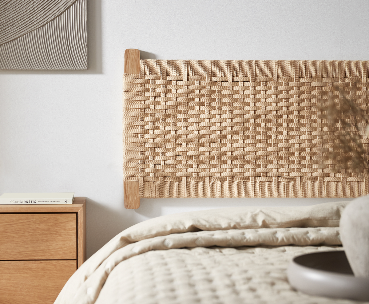 Nærbillede af et soveværelse med en vævet naturfarvet sengegavl i træ, beige sengetøj og et lys eget natbord med en bog med titlen SCANDI RUSTIC; lys og luftig skandinavisk stemning.