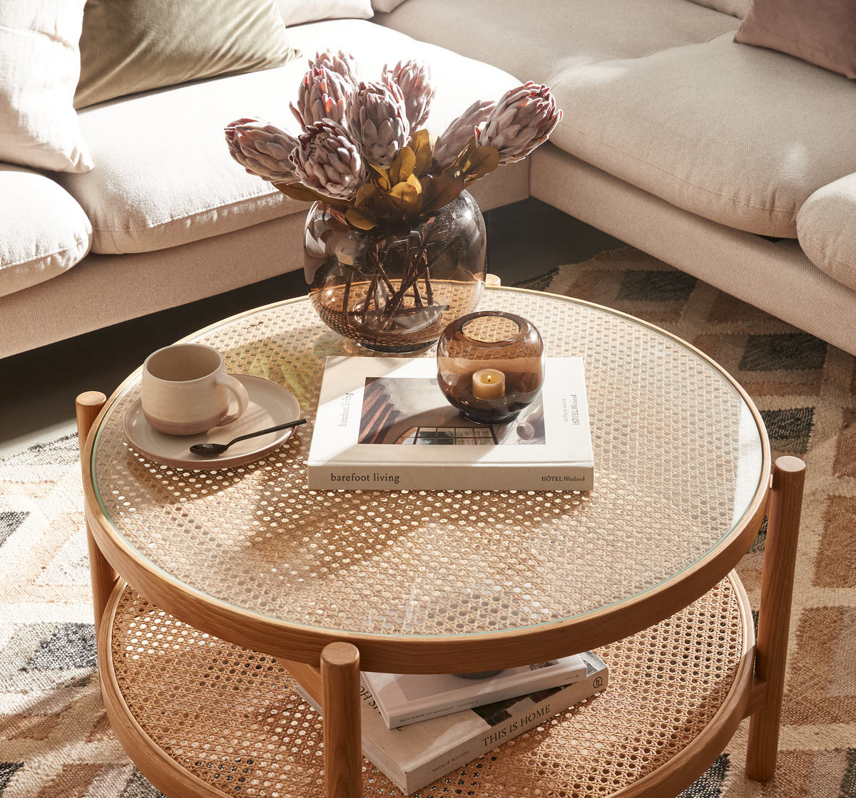 Rundt rattankaffebord med glasplade i en stue, stylet med brun vase med protea, et lys i lille glaslygte, en kop på underkop og bøgerne “barefoot living” og “This Is Home”.