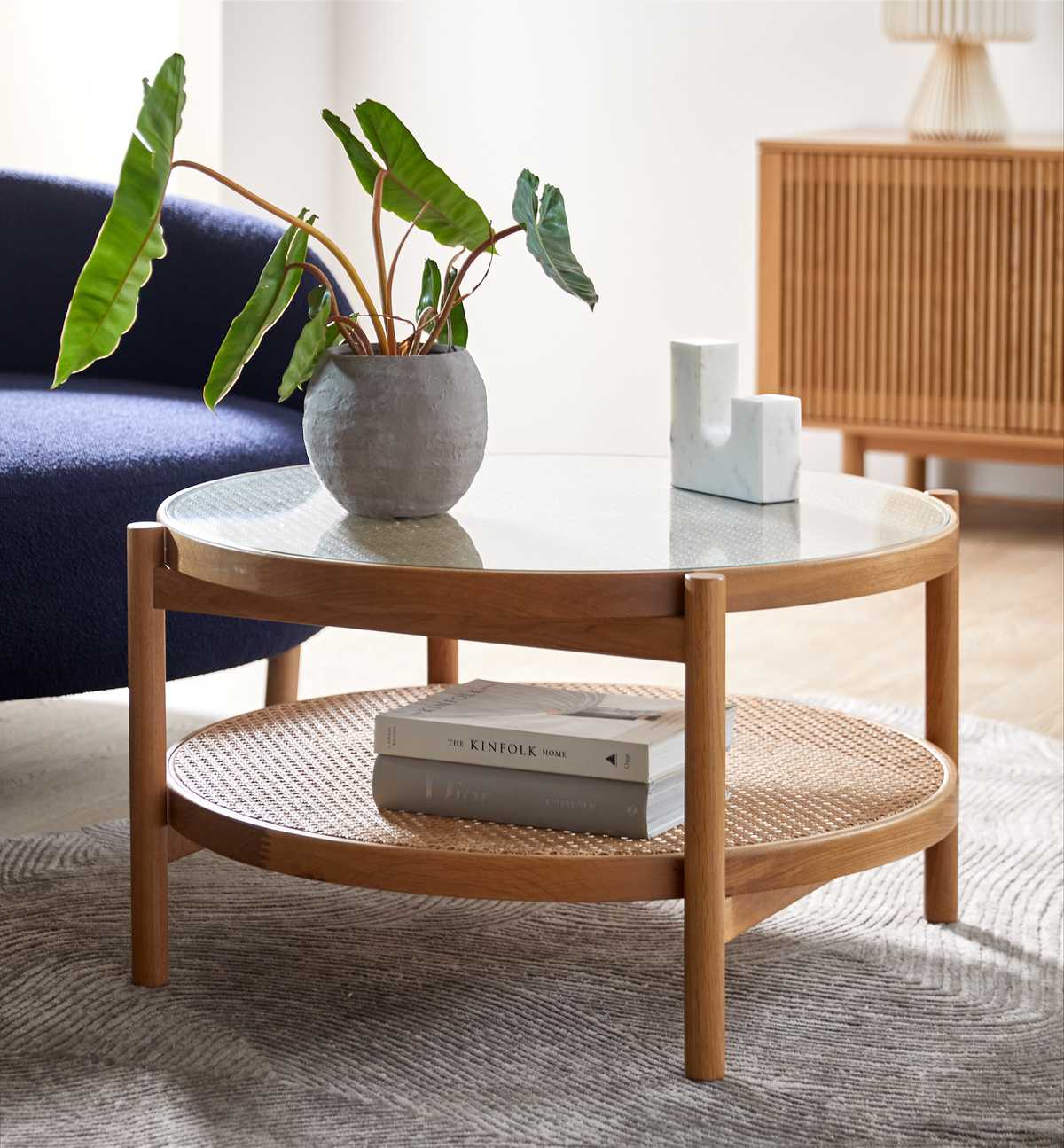 Rundt sofabord i træ med glasplade og rattanhylde; potteplante, marmordekoration og en bog med titlen The Kinfolk Home på den nederste hylde i en lys stue.