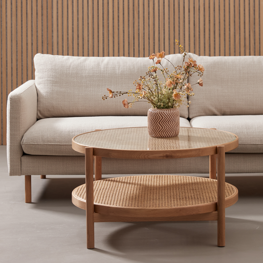 Beige sofa med rundt sofabord i træ med glasplade og rattanhylde; vase med tørrede blomster foran en lamelvæg i træ.