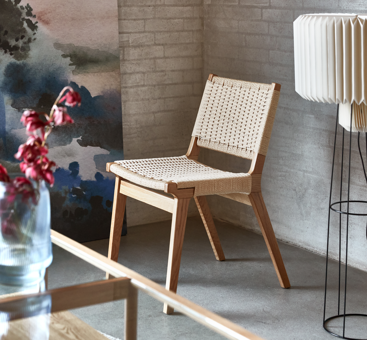 Lys egetræsstol med flettet sæde og ryg i en lys stue, mod lys murstensvæg, med glasvase med røde blomster, abstrakt vægkunst og hvid plisseret gulvlampe.