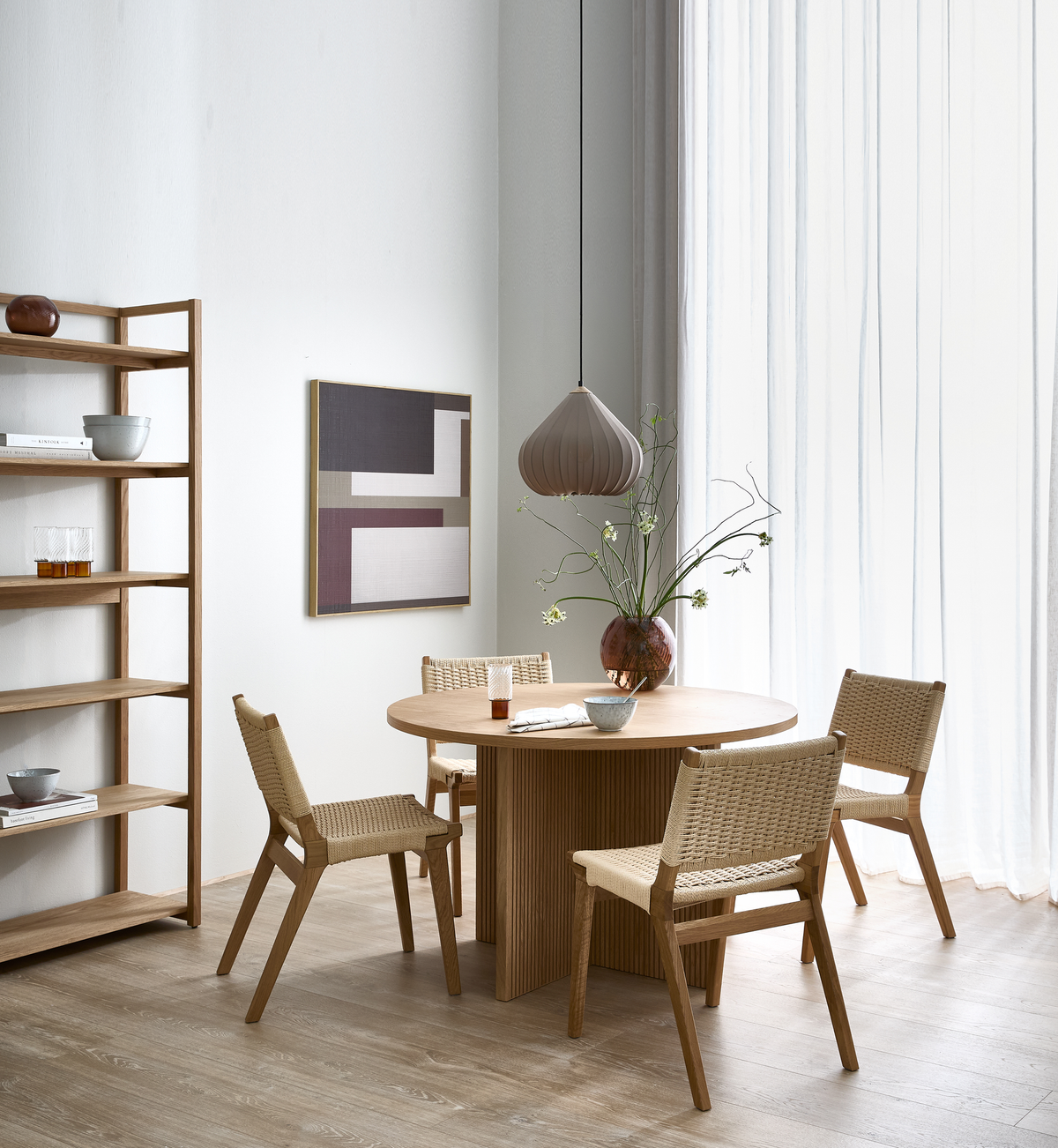 Lyst spisestue med rundt spisebord i træ, fire fletstole, hængende pendel og en vase med grønne grene ved stort vindue med lette gardiner.