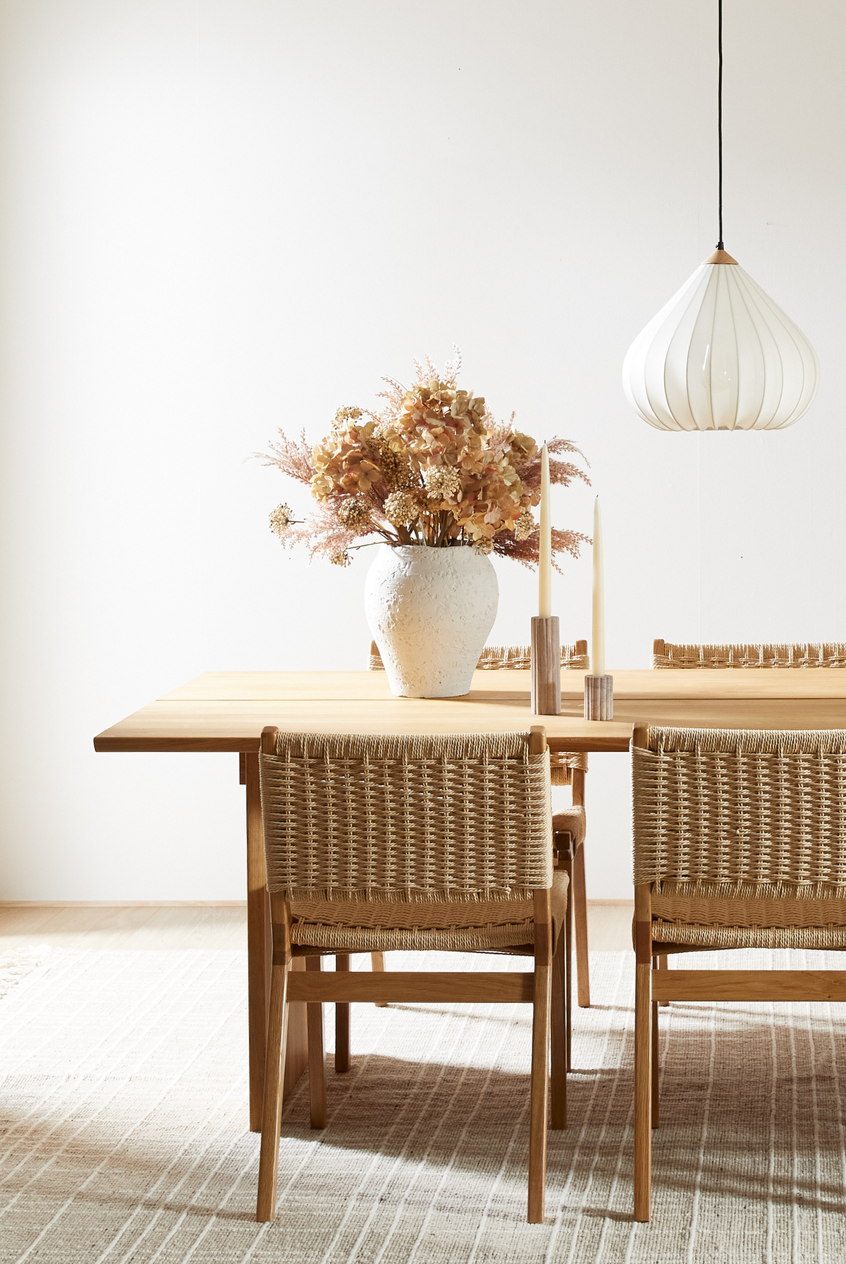 Spiseplads med lyst træbord, fletstole, hvid pendellampe, stor hvid vase med tørrede blomster og to slanke lys på et neutralt tæppe.