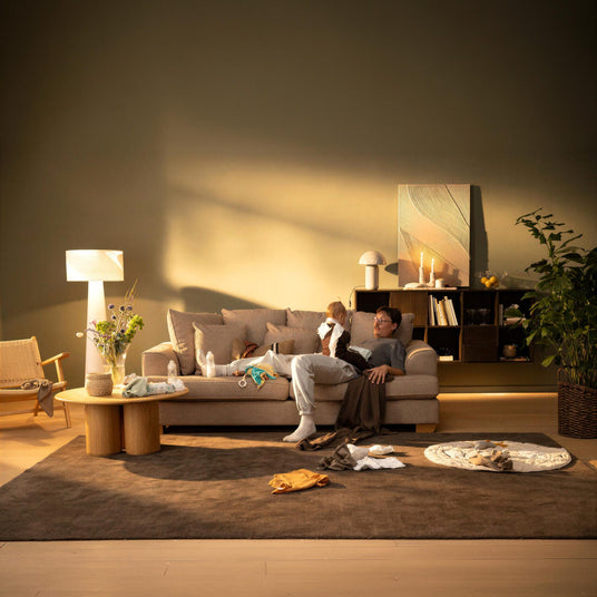 En forælder sidder i en beige sofa med en baby i en varmt oplyst stue; babytøj og tilbehør ligger spredt på et brunt tæppe, med gulvlampe, blomster i vase, tændte lys og en stor grøn plante i baggrunden.