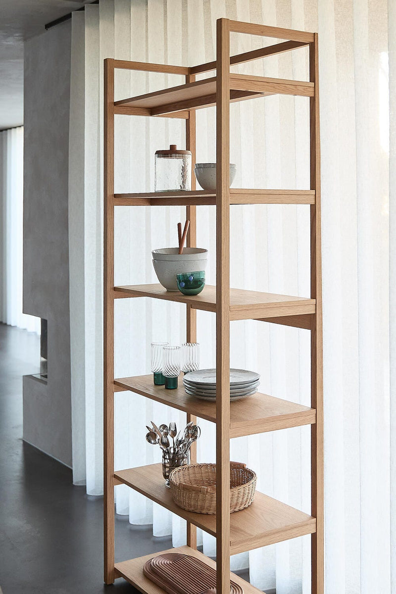 Åben reol i lyst træ med skåle, glas, tallerkener og flettekurv i en lys spisestue med lette gardiner.