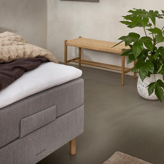 Roligt soveværelseshjørne med grå polstret seng med “nocturne”-mærke, beige plaid, flettet træbænk og grøn potteplante i hvid krukke på neutralt gulv.