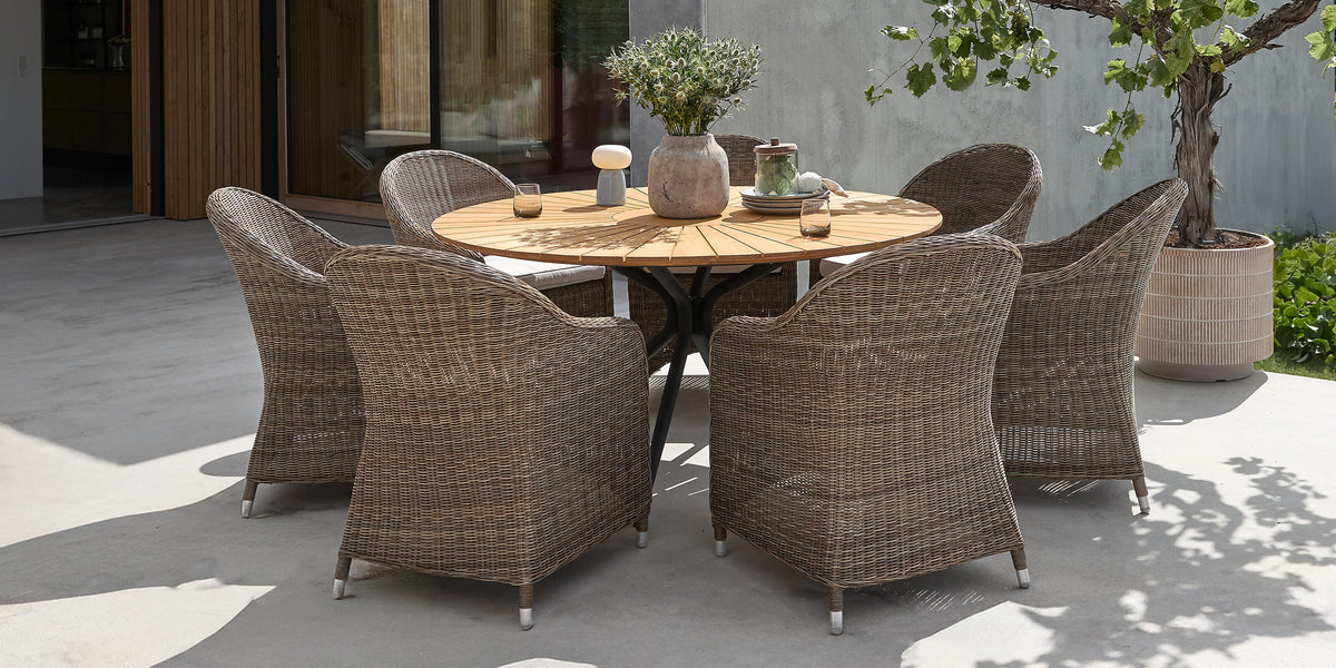 Moderne terrasse med rundt træbord og seks fletstole ved beton- og træhus, krukket træ og dagslys.