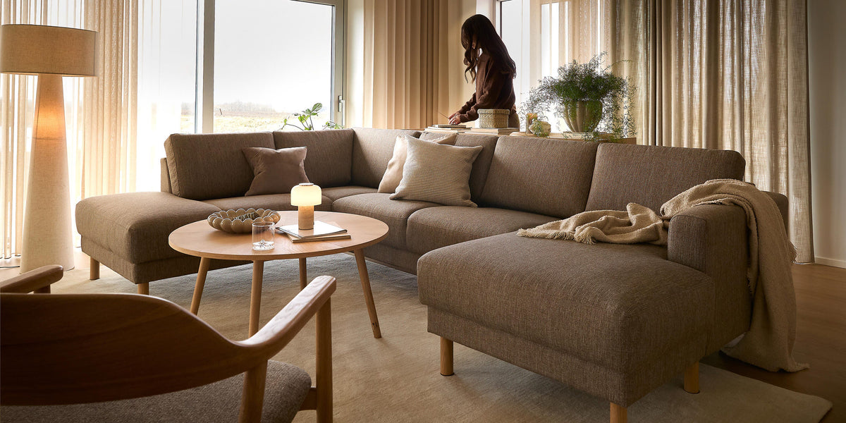 Lyst stue med beige gardiner, brun hjørnesofa i stof, rundt træbord med lille lampe og plaid på chaiselongen; en person står ved vinduet.