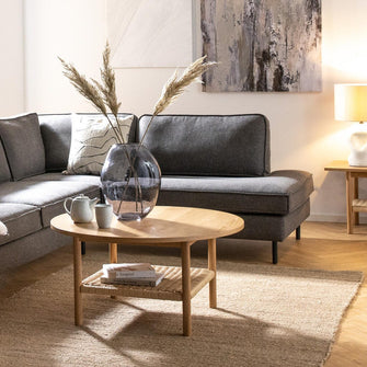 Lys skandinavisk stue med grå hjørnesofa, rundt sofabord i træ, glasvase med tørrede strå, neutral jutemåtte og to abstrakte billeder; pendel og bordlampe skaber en varm, luftig stemning.