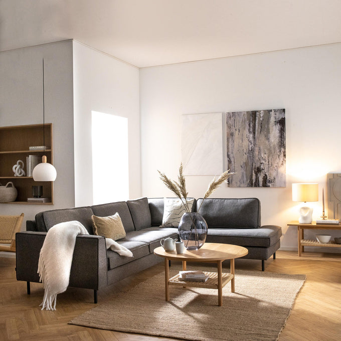Lys skandinavisk stue med grå hjørnesofa, rundt sofabord i træ, glasvase med tørrede strå, neutral jutemåtte og to abstrakte billeder; pendel og bordlampe skaber en varm, luftig stemning.