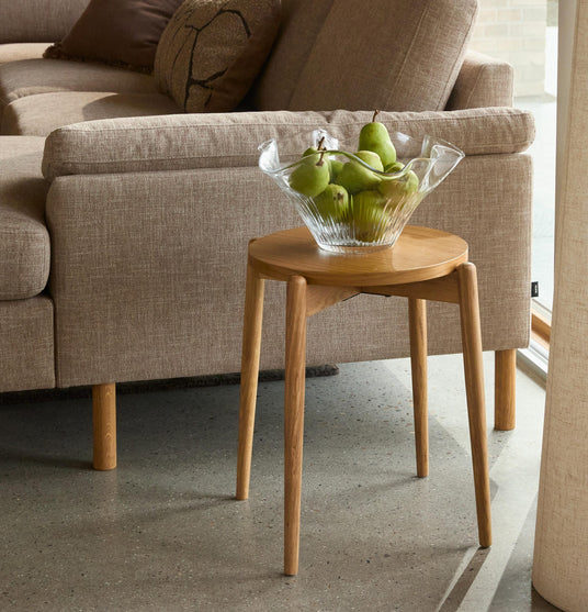 Beige sofa med lille rundt træ-sidebord og glasskål med grønne pærer i en lys stue.