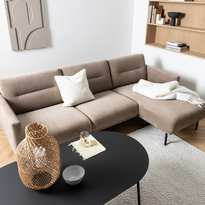 Beige hjørnesofa med hvid pude og plaid, sort sofabord med flettet lanterne, skål og glas; lys tæppe og træhylde på væggen i en lys stue.