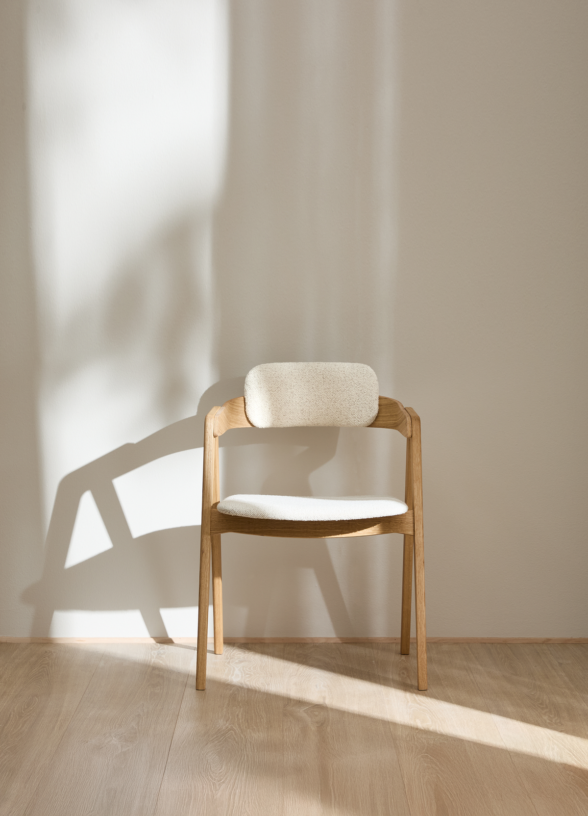 Lys træstol med polstret sæde og ryglæn i blødt sollys mod beige væg.