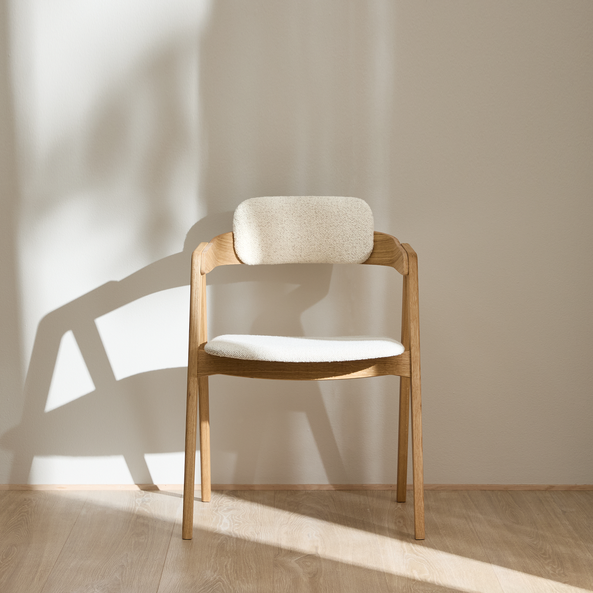 Lys træstol med lys polstret sæde og ryglæn mod beige væg i blødt dagslys.