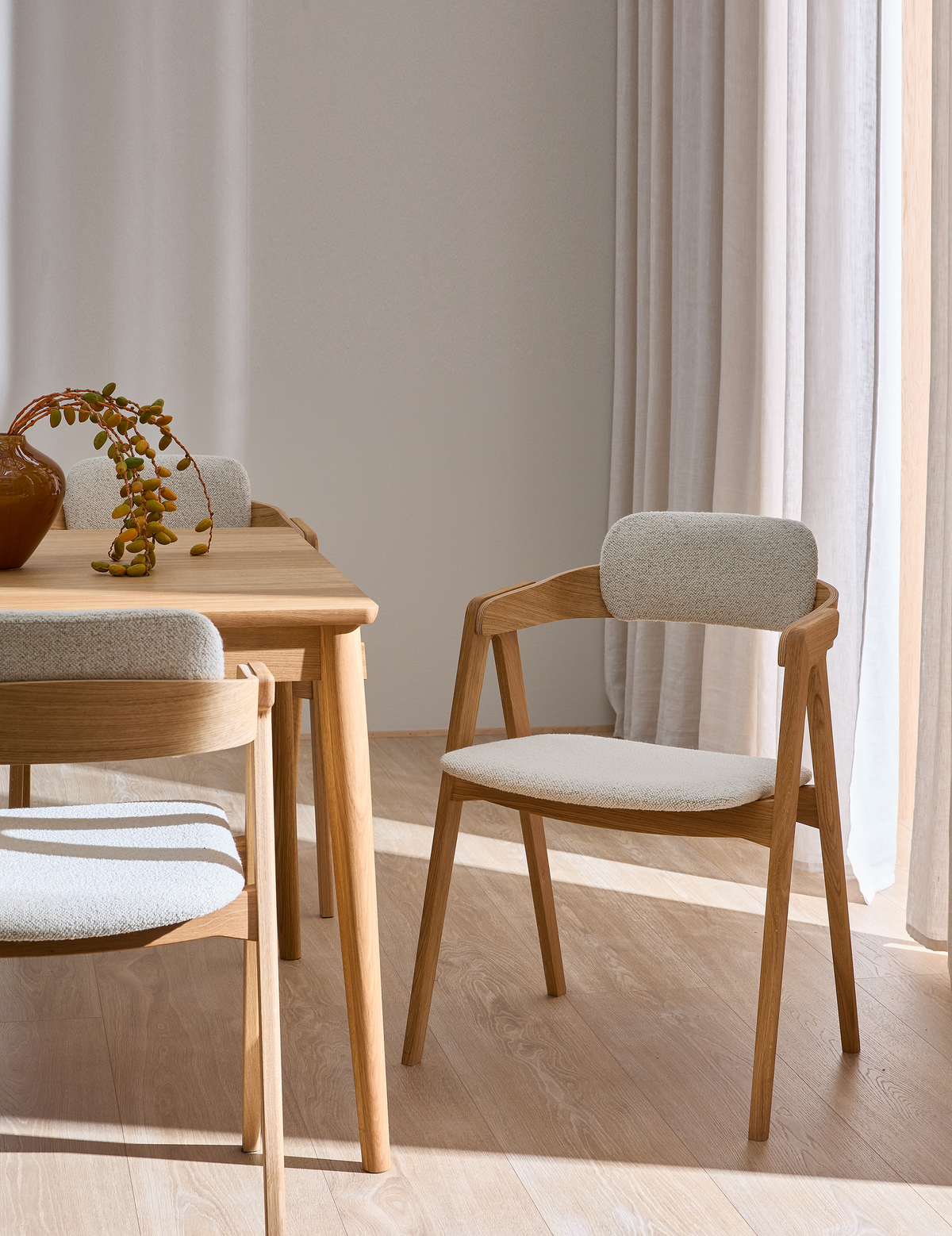 Lyst spiserum med egetræsbord og beige polstrede stole, solstrejf gennem lyse gardiner og en vase med hængende grøn gren.