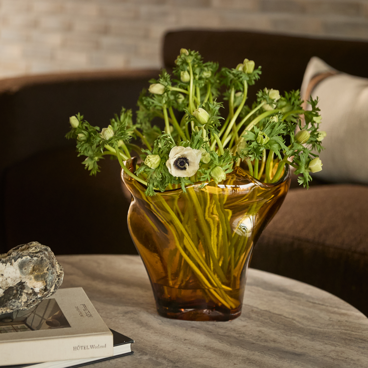 Ravgul glasvase med hvide anemoner på et rundt stenbord ved siden af en sten og nogle bøger, hvoraf en har titlen ”HOTEL Weekend”, i en hyggelig stue.