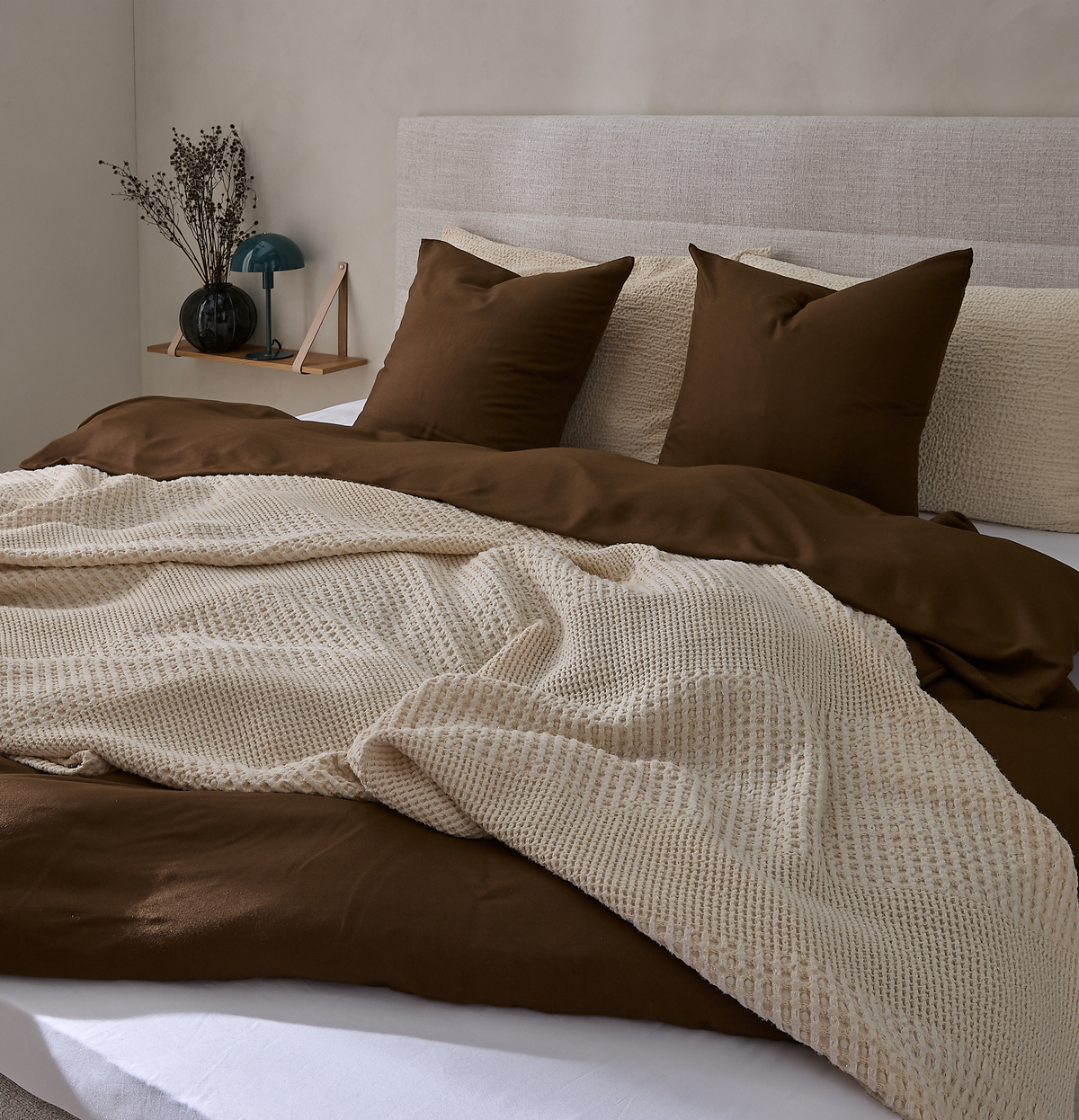 Redt seng med brunt dynebetræk, beige strikket plaid og matchende puder i et neutralt soveværelse med polstret sengegavl og lille hylde med vase og lampe.