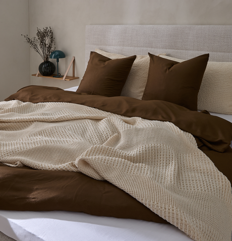 Redt seng med brunt dynebetræk, beige strikket plaid og matchende puder i et neutralt soveværelse med polstret sengegavl og lille hylde med vase og lampe.
